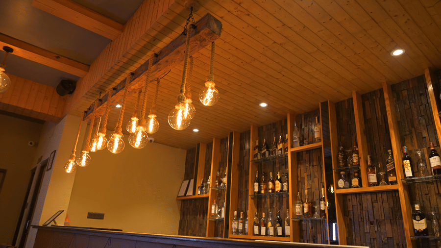 Bar counter at Hotel Sonar Bangla, Sundarban with hanging Edison bulbs on rope, wooden-clad ceiling, and shelves displaying assorted liquor bottles.