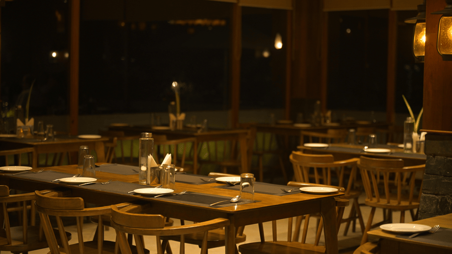Restaurant at Hotel Sonar Bangla, Sundarban at night with set wooden tables, spindle-back chairs, pendant lighting, and a wall-mounted lantern fixture.