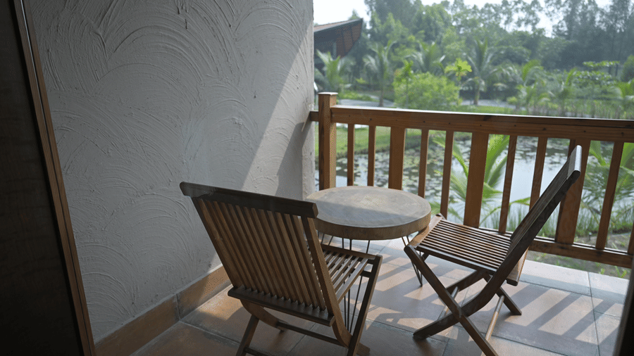 A balcony with 2 wooden chairs and a small round table overlooking trees at Hotel Sonar Bangla, Sundarban.