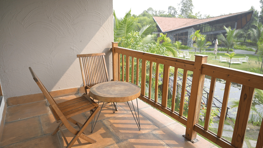 A tiled balcony with 2 wooden chairs and a round table overlooking green trees and a building at Hotel Sonar Bangla, Sundarban.