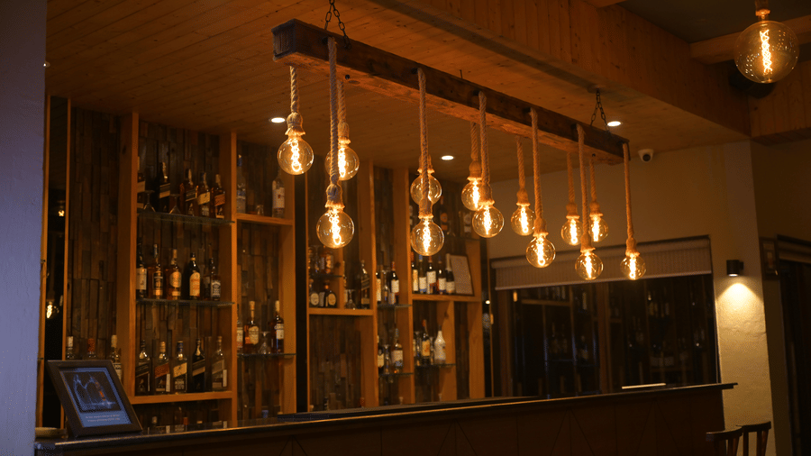 A bar counter featuring a row of hanging warm lights and shelves stocked with bottles at Hotel Sonar Bangla, Sundarban.