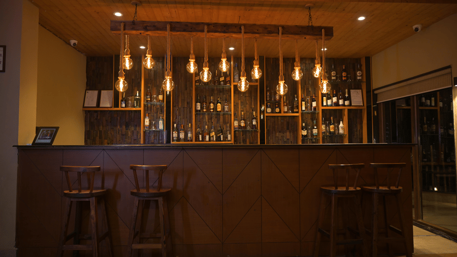 Frontal view of a dark wooden bar counter with high stools and a row of pendant lights at Hotel Sonar Bangla, Sundarban.