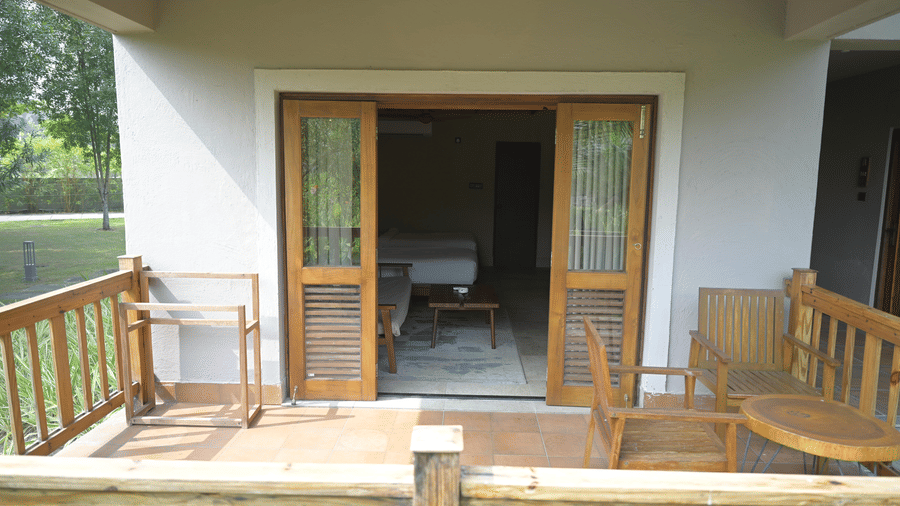 A wooden balcony with chairs and a small table looking through glass doors into a hotel room at Hotel Sonar Bangla, Sundarban.