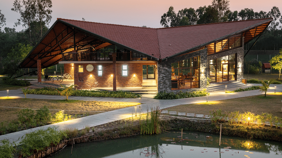 Hotel Sonar Bangla, Sundarban building at twilight with slanting roof, brick and stone facade, lit pathways, lily pond, and bicycles parked outside.