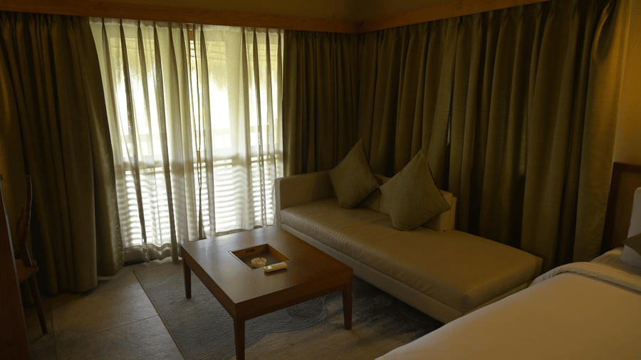 Lounge area in a hotel room with a beige sofa, wooden coffee table, and olive green curtains at Hotel Sonar Bangla, Sundarban. 