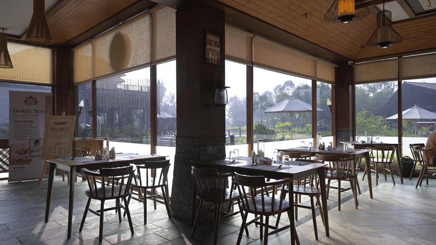 Indoor cafe at Hotel Sonar Bangla, Sundarban with wooden tables, chairs, and floor-to-ceiling windows overlooking the gardens.