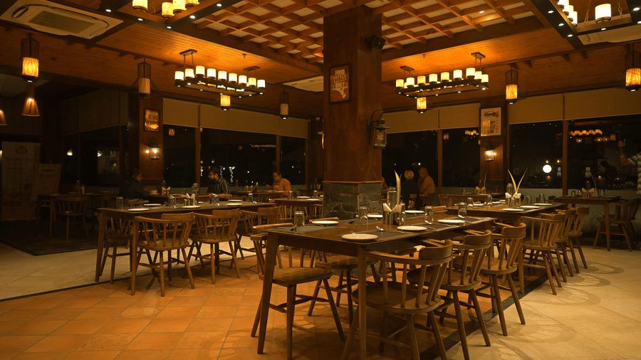Warmly lit restaurant at Hotel Sonar Bangla, Sundarban with wooden coffered ceiling, lantern pendants, long dining tables, and central stone-clad pillar.