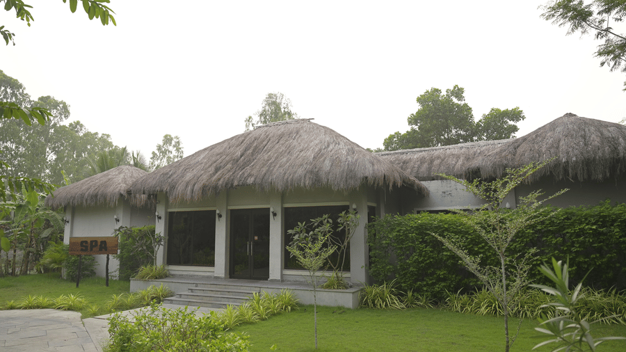 Wide shot of a thatched-roof building surrounded by green plants and a lawn at Hotel Sonar Bangla, Sundarban.