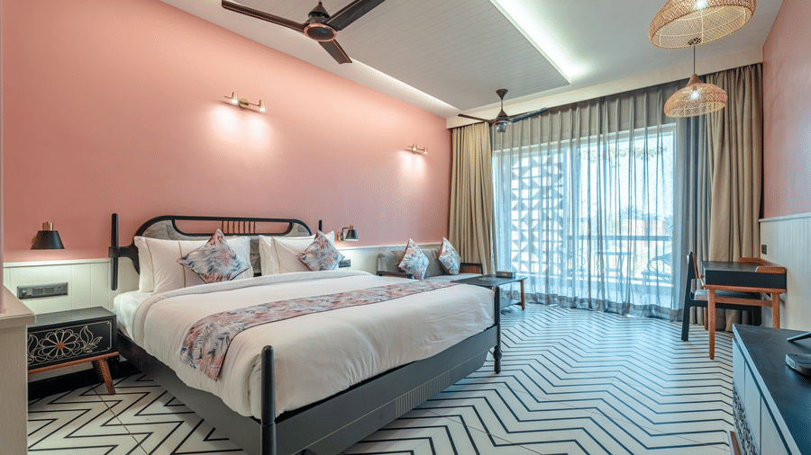 A bright Deluxe Room with large glass balcony doors allowing natural sunlight into the bedroom, complemented by modern interiors and soft pink walls | Hotel Sonar Bangla, Susunia