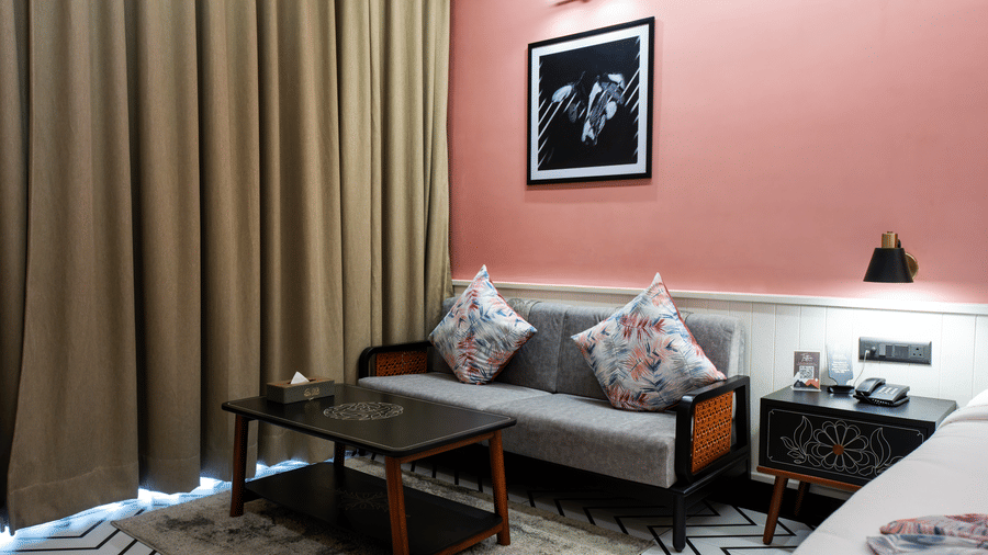 Our cosy Susunia room corner with a sofa seating area, wooden coffee table, long curtains, and warm pink accent walls beside the bed | Hotel Sonar Bangla, Susunia