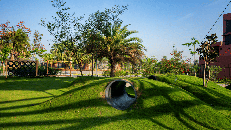 A lush green garden lawn featuring tunnel play structures, palm trees, and long evening shadows spread across the landscaped outdoor area | Hotel Sonar Bangla, Susunia