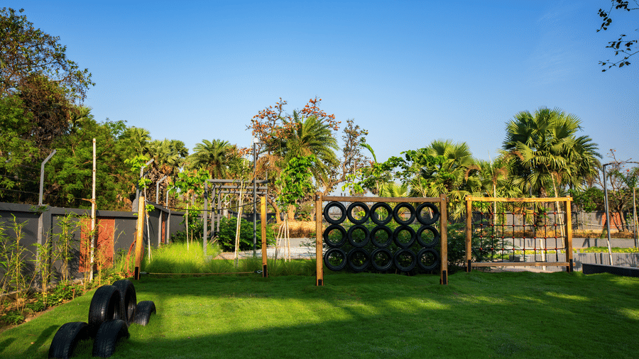 A landscaped green lawn with outdoor play elements, decorative fencing, and palm trees spread across the sunny garden area of the resort | Hotel Sonar Bangla, Susunia