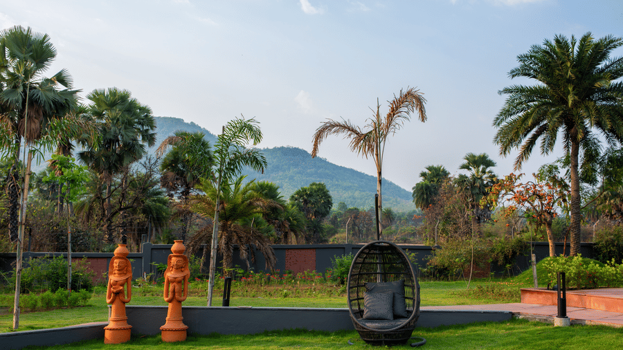 A landscaped garden area featuring artistic sculptures, palm trees, and open green lawns under a bright daytime sky | Hotel Sonar Bangla, Susunia