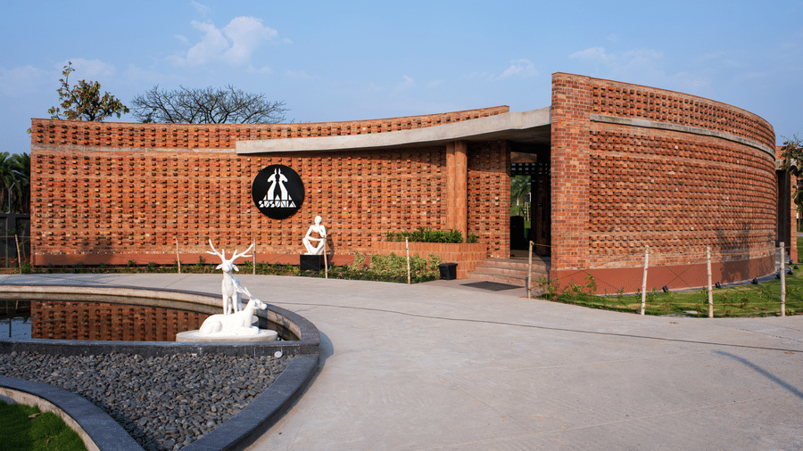 The curved brick entrance of the reception with landscaped pathways and a decorative fountain at the driveway entrance under a clear blue sky | Hotel Sonar Bangla, Susunia