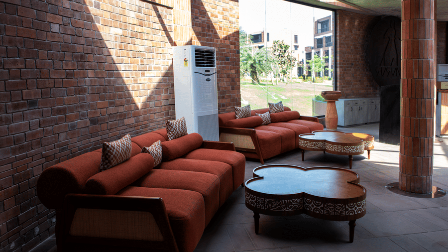 A cosy indoor lounge area with red cushioned sofas, round coffee tables, and natural sunlight streaming through the open brick-walled seating space | Hotel Sonar Bangla, Susunia