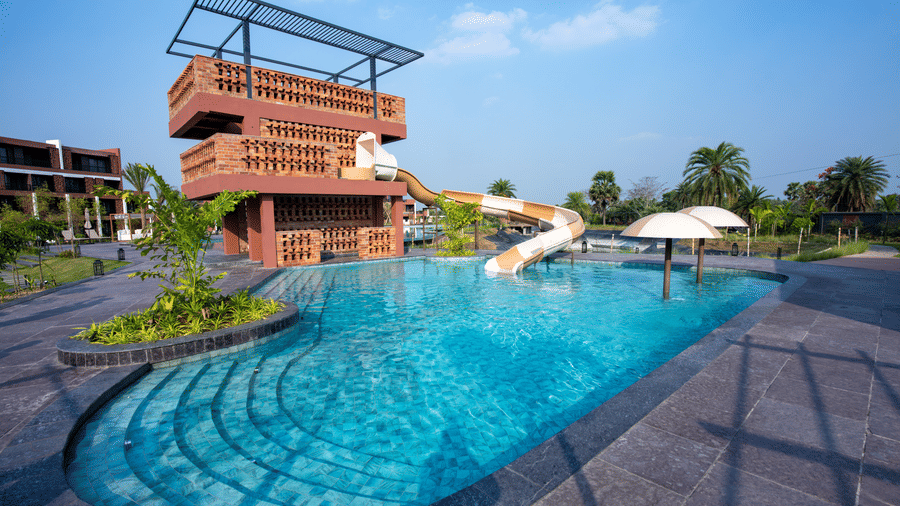 The curved outdoor swimming pool with clear blue water, surrounding deck space, a slide for kids, and the resort building overlooking the pool area | Hotel Sonar Bangla, Susunia
