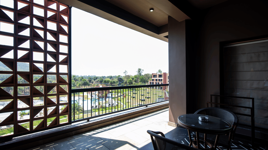 A private balcony seating area with outdoor chairs and a table overlooking the landscaped resort grounds through decorative lattice-style walls | Hotel Sonar Bangla, Susunia