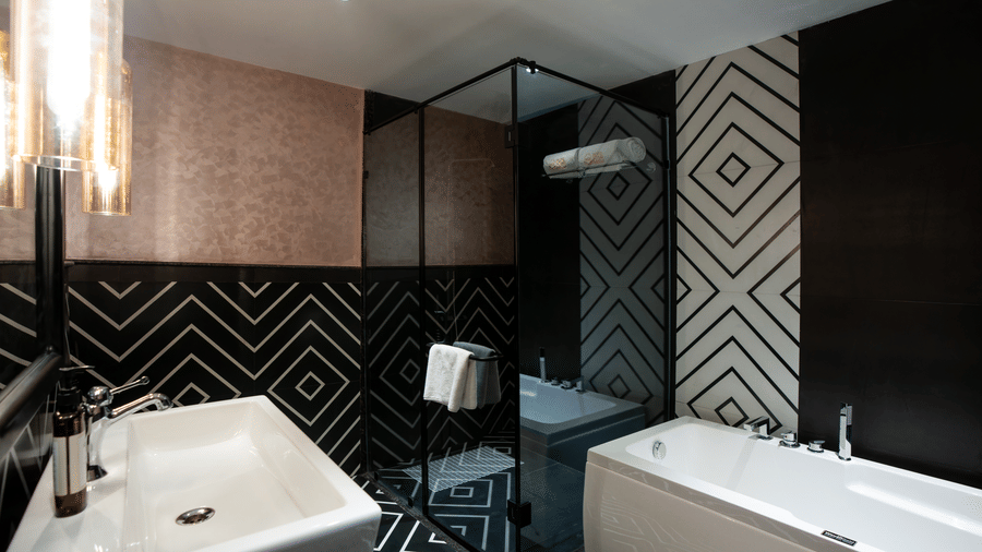 A stylish suite bathroom with geometric black-and-white wall patterns, a bathtub, vanity counter, and glass shower enclosure | Hotel Sonar Bangla, Susunia