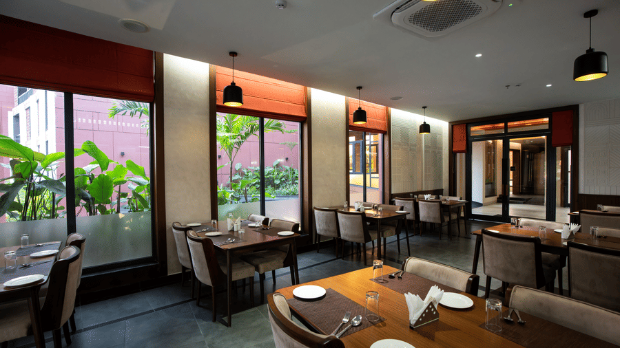 Indoor seating at Tattva Restaurant with wooden tables, cream upholstered chairs, warm pendant lights, and large windows overlooking lush greenery | Hotel Sonar Bangla, Susunia