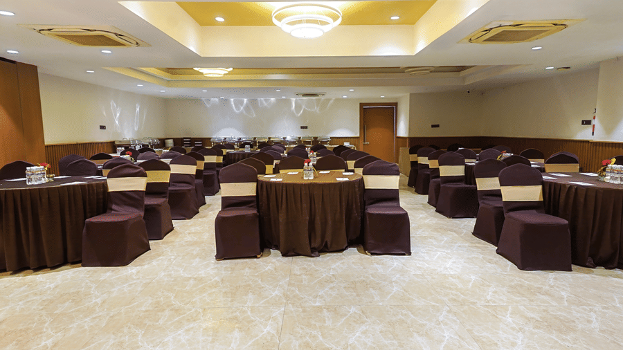 Wide shot of Ganga hall with a white ceiling adorned with circular lights along with 2 air-conditioners and chairs and tables arranged in rows at Hotel Sonar Bangla, Taki.