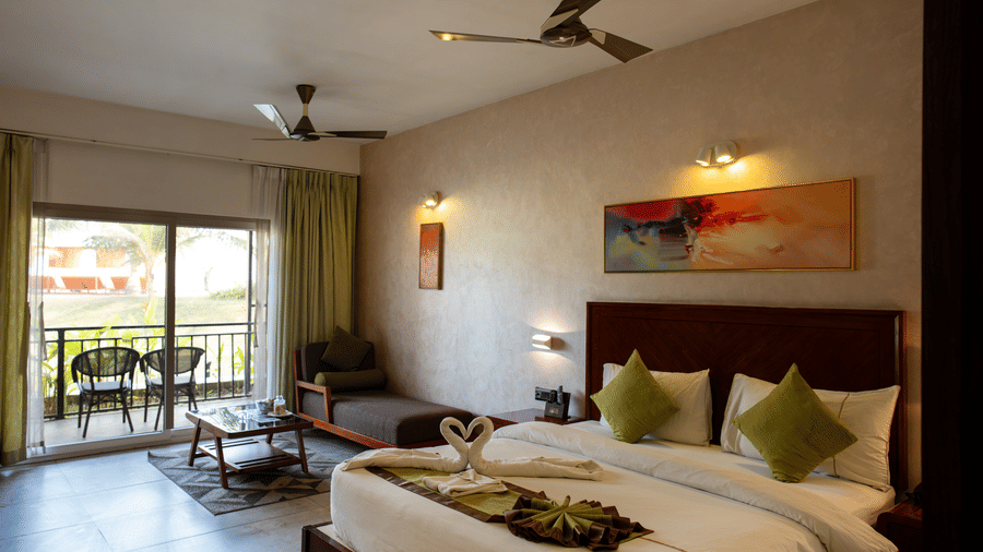 Room featuring green curtains open to a balcony view with 2 chairs kept outside, a large bed, 2 fans, a painting above the bed, and swan shaped towel pair kept at the foot of the bed at Hotel Sonar Bangla, Taki.