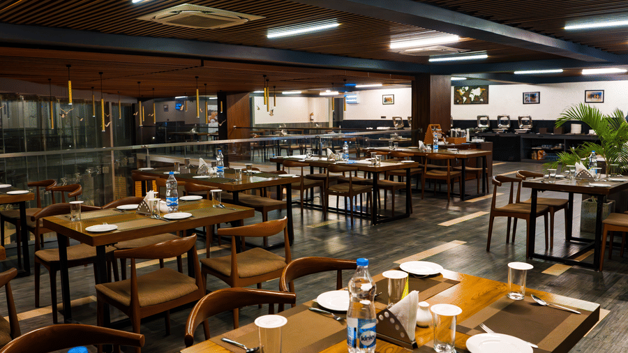 A restaurant featuring a wooden roof and 5 tables with seating for 6 members each with plates and water bottles arranged on it at Hotel Sonar Bangla, Taki.