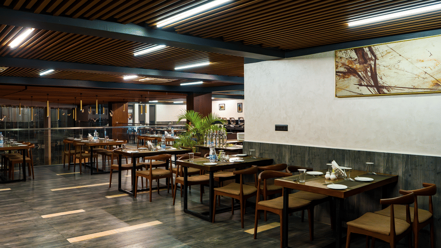 Side angle view of a restaurant featuring a wooden roof and 5 tables with seating for 6 members each with plates and water bottles arranged on it at Hotel Sonar Bangla, Taki.