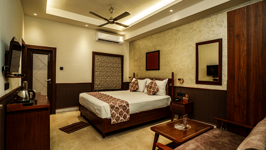 A hotel bedroom with a double bed, a sofa, warm ceiling lights, a TV, an AC unit, a fan, and wooden furniture | Hotel Sonar Bangla, Tarapith