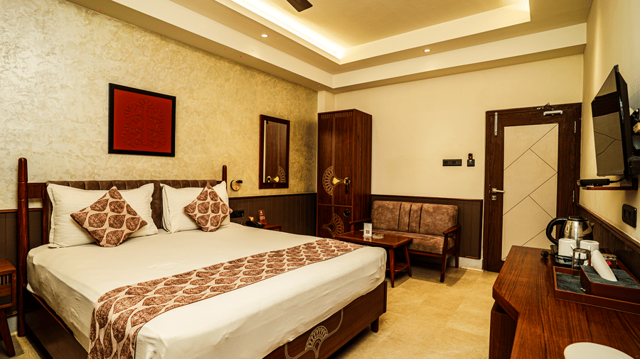 The Deluxe Room with a large bed, bedside lighting, wooden decor, a TV, a tea/coffee maker, a wardrobe, and a sofa | Hotel Sonar Bangla, Tarapith