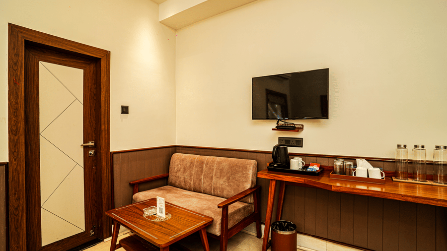 The seating area in our Deluxe Four Bedded Room featuring a cushioned chair, a coffee table, a wall-mounted television, and a table with water bottles | Hotel Sonar Bangla, Tarapith