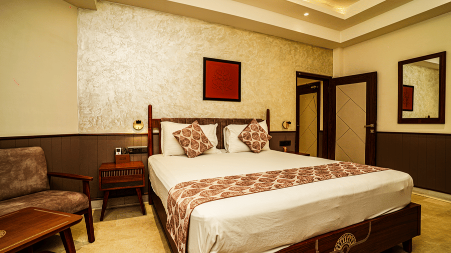 A bedroom with a double bed, a cosy chair, a coffee table, wooden decor, and a night stand in our Deluxe Suite | Hotel Sonar Bangla, Tarapith