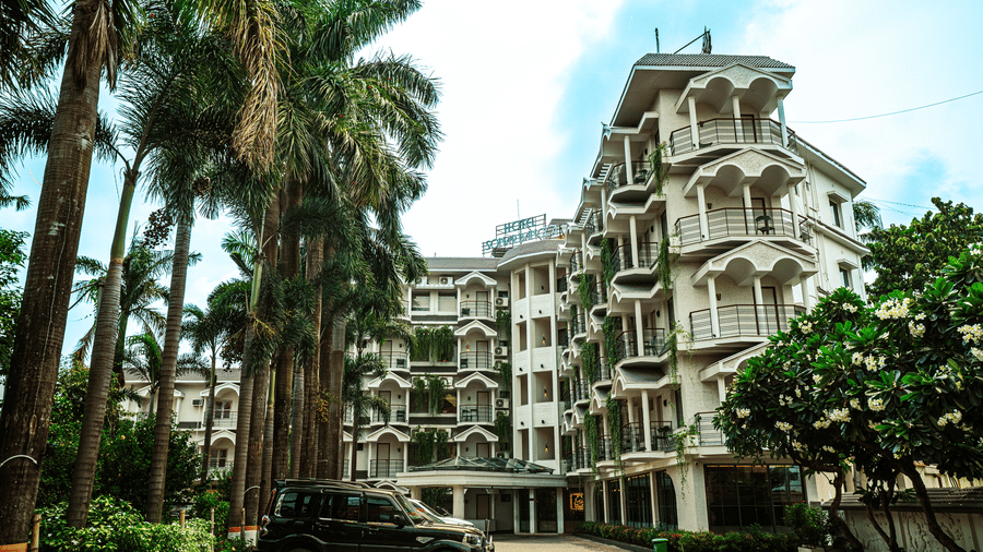 Exterior view of Hotel Sonar Bangla, Tarapith, featuring multi-storey architecture, tall palm trees, parked vehicles, and landscaped surroundings.