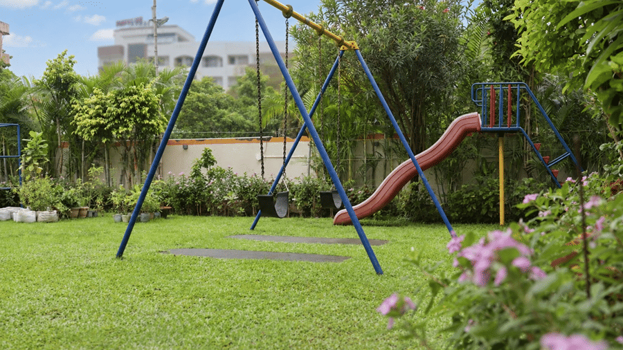 An outdoor play area at Hotel Sonar Bangla, Tarapith featuring a swing set, metal slide, manicured lawn, flowers, and surrounding greenery.