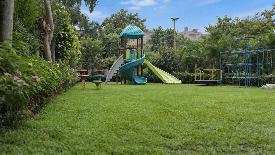 An outdoor play area at Hotel Sonar Bangla, Tarapith featuring spiral slides, play tower, climbing structure, flower borders, and open lawn.