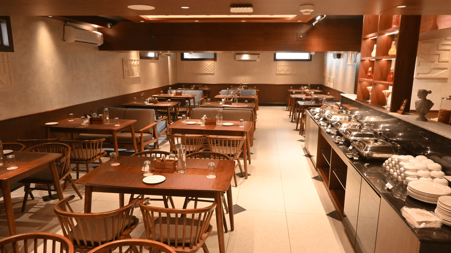 A restaurant dining room set up for a buffet, featuring rows of empty wooden tables with woven cane chairs, a long serving counter with covered chafing dishes on the right, and warm wooden panelling and recessed lighting throughout at Hotel Sonar Bangla, P