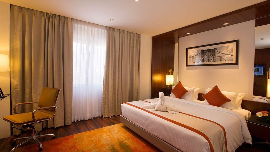 Another view of a hotel room with a large bed with white and orange linens, a brown office chair and sheer curtains over a window - Hotel Southend by TGI - Bommasandra, Bangalore