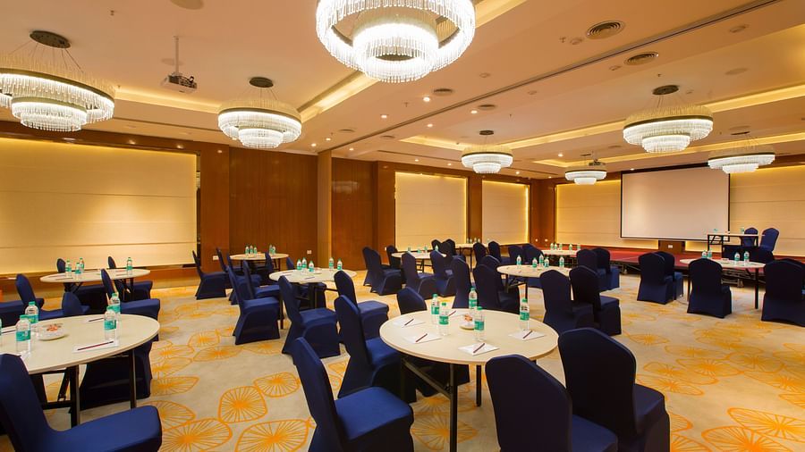 A large conference hall with circular tables, blue chairs and a projector screen, ready for a seminar - Hotel Southend by TGI - Bommasandra, Bangalore