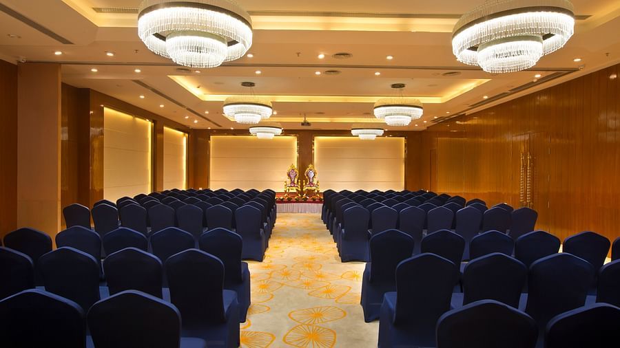 A spacious conference hall with rows of blue chairs facing a stage with golden figurines, illuminated by large chandeliers - Hotel Southend by TGI - Bommasandra, Bangalore