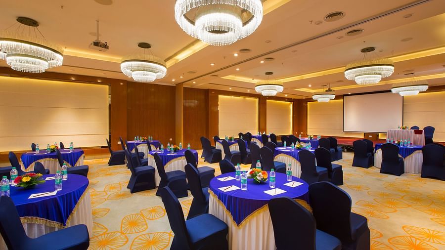 Another view of a large conference hall with circular tables, blue chairs and a projector screen, ready for a seminar - Hotel Southend by TGI - Bommasandra, Bangalore