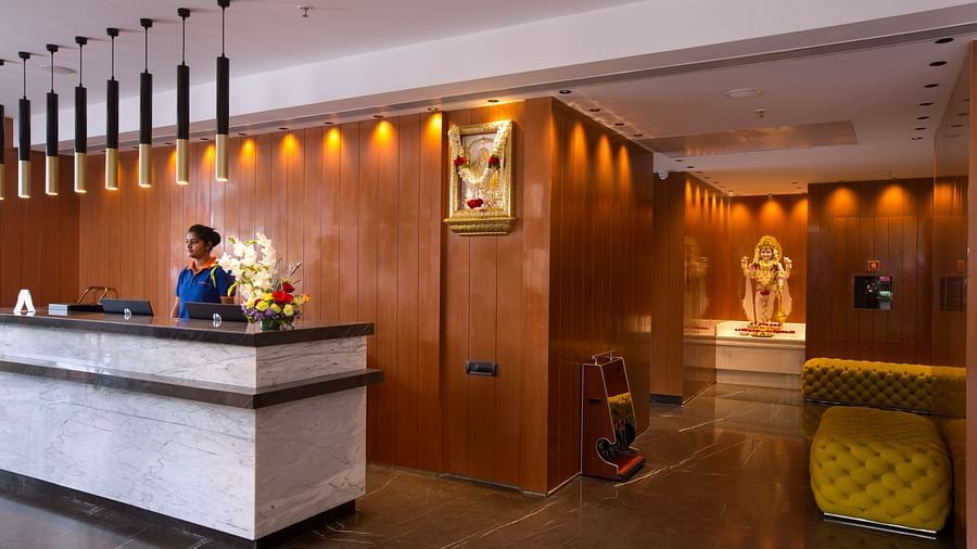 The hotel reception desk with a wooden backdrop and a flower arrangement, flanked by a statue of god in a glass case - Hotel Southend by TGI - Bommasandra, Bangalore