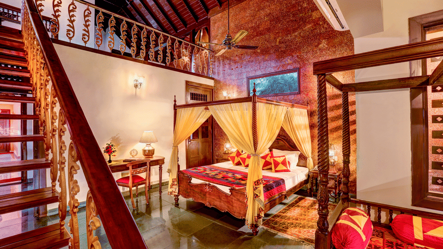 An overview of the Malnad Attic Suite with a bedroom having two poster beds and a wooden stair leading to the attic of the room at Hoysala Village Resort, Hassan.