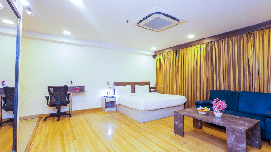A bright hotel room with a large bed, wooden floors, a blue sofa, and a desk with a chair - Hyphen Business Hotel, Noida
