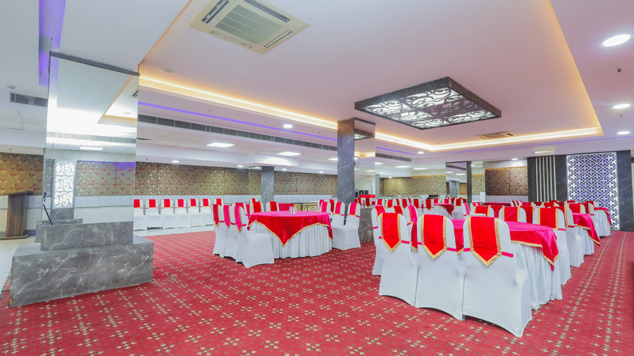 A wide shot of a banquet hall with multiple round tables arranged for an event, featuring red and white decor - Hyphen Business Hotel, Noida