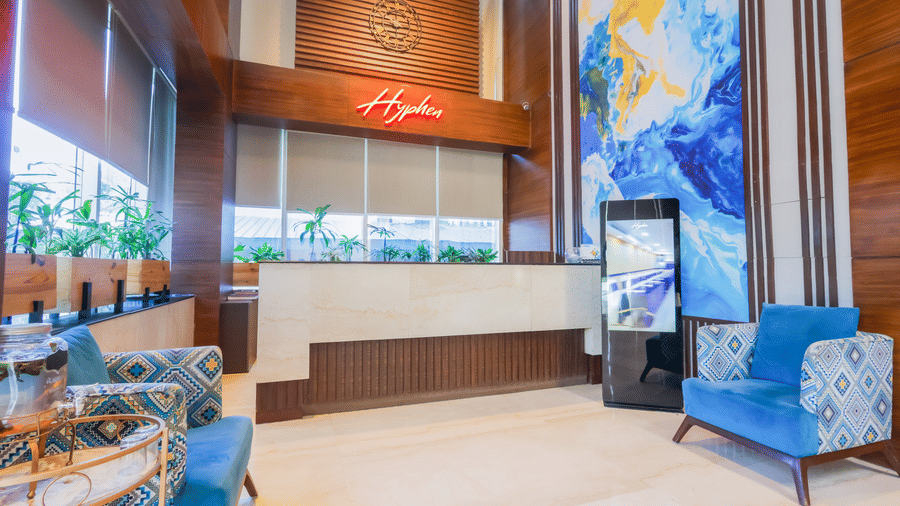 A hotel lounge area with comfortable blue armchairs, wooden accents, and decorative elements - Hyphen Business Hotel, Noida