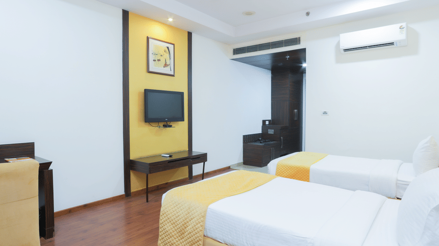 Bright twin-bedroom with wooden furniture, wall-mounted TV, and white walls at Hyphen Premier Hotel, Meerut.
