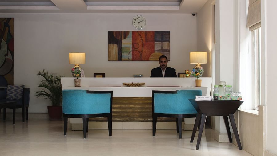 An image of a reception lobby featuring a reception desk where a reception is standing and in front of them there are two chairs placed along with a table at the corner of the room - Inde Hotels Vista Woods