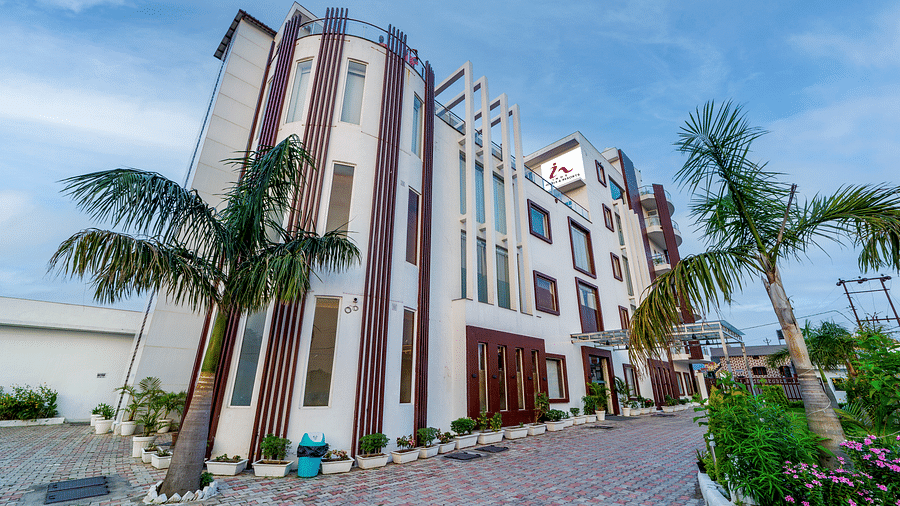Exterior view of Inde Reva Resorts Rishhikesh showing the multi-storey building with palm trees, paved driveway, and lush green on the right.