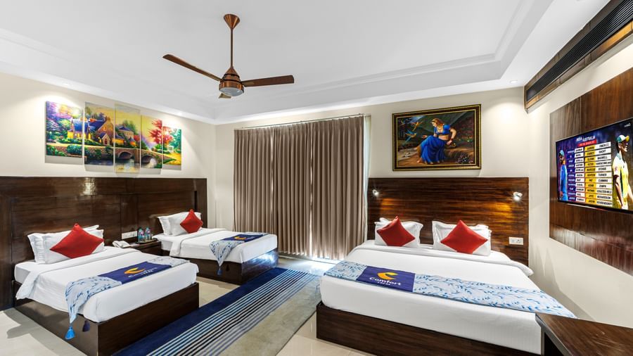 Well furnished huge room with a king size bed and twin beds featuring a ceiling fan, artwork above bedside table, and TV unit opposite the beds at Inder Reva Resorts Rishhikesh.