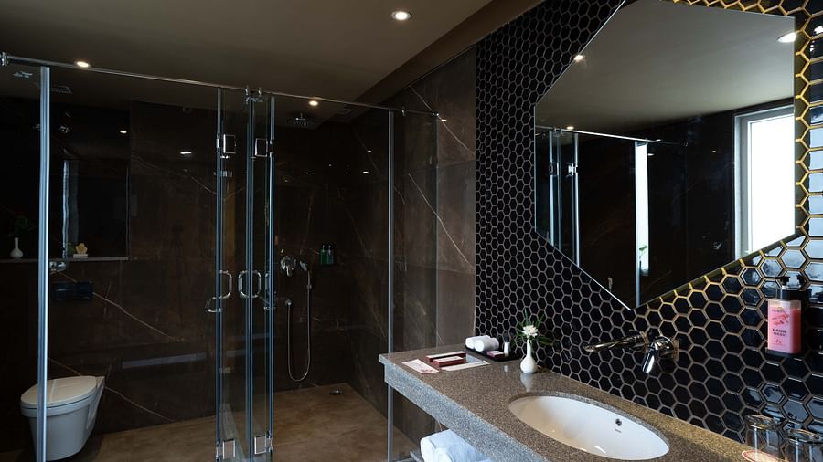 Hexagon-shaped mirror above the washbasin with transparent glass divider separating the showering and toilet areas in the Deluxe room's bathroom at Inde Signature Jaipur.