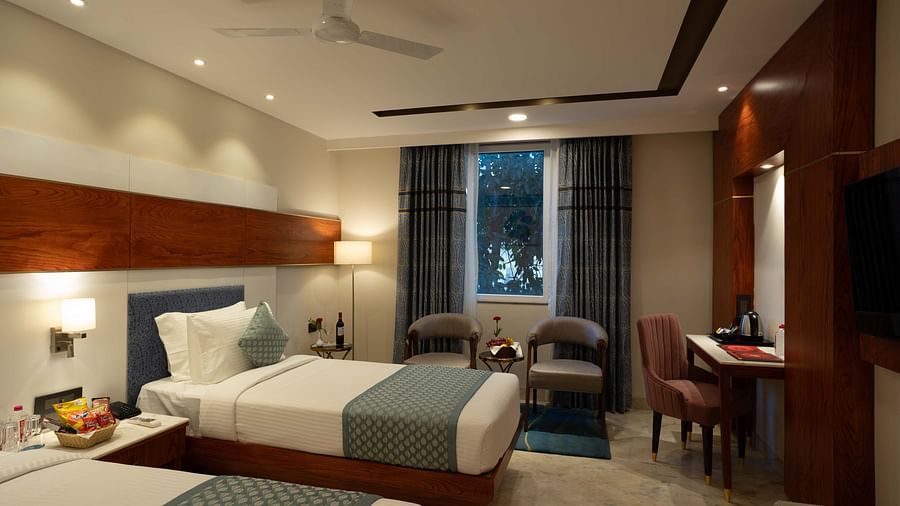 Deluxe Room with twin beds along with a wooden side table with a lamp, wooden table and chair, and two cushioned pieces of furniture at Inde Signature Jaipur